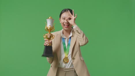 asian business woman in a suit with a gold medal and trophy showing okay hand sign over eye and smiling to camera as the first winner on green screen background in the studio