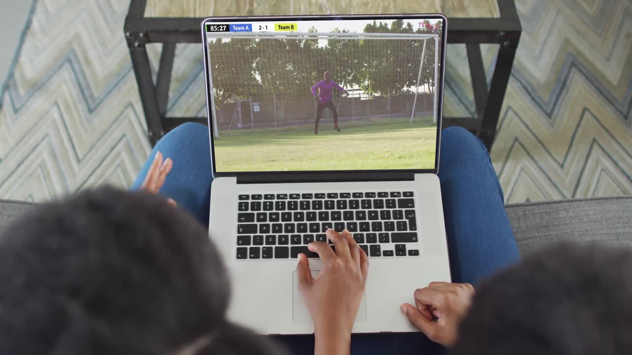 video de dos personas afroamericanas sentadas en el sofá y viendo un partido de fútbol en una computadora portátil
