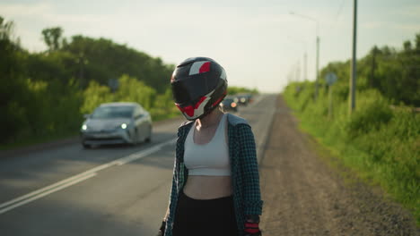 a woman wearing a motorcycle helmet stands on the roadside, giving a thumbs-up as cars pass by in the opposite direction, she is dressed in a plaid shirt and crop top