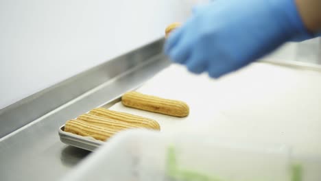 Unrecognizable-female-confectioner-hands-in-blue-gloves-lay-out-eclairs-in-threes-in-a-row-on-baking-tray-with-baking-parchment-underneath