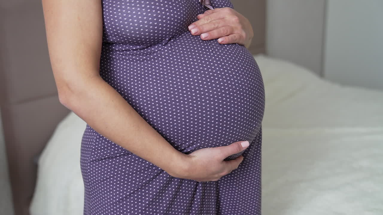 Midsection of pregnant woman wearing purple night dress and caressing her big belly. Expecting mother standing by the large bed in her cozy bedroom Close up.