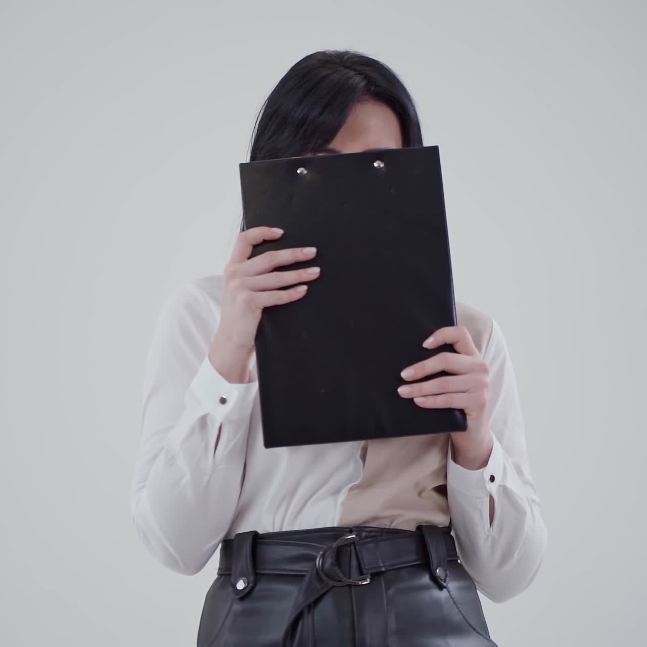 Portrait of a beautiful brunette woman. Attractive lady in white blouse holding black folder and closing her smiling face on the light background. Slow motion.