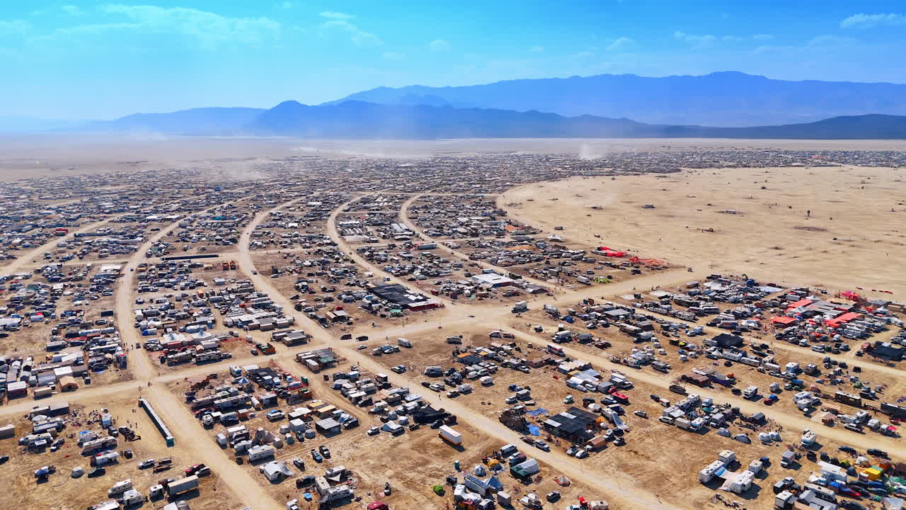 Wide aerial of curved desert city. Endless rows of camps and streets form a sweeping arc across the open desert plain