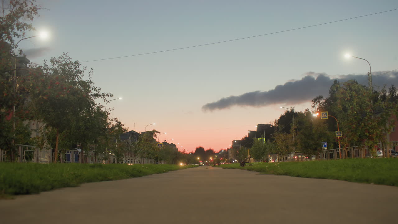 Vibrant urban evening view with illuminated street lights, distant moving cars, aligned trees, residential buildings, and dramatic sunset sky creating serene atmosphere of cityscape at dusk