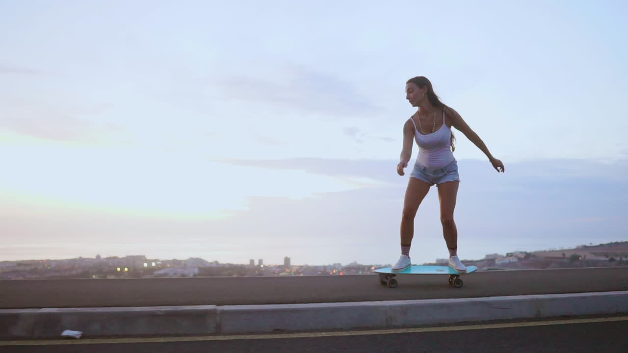 As the sun sets, a gorgeous and stylish young skateboarder rides her board along a mountain road, wearing shorts and showcasing the mountains' spectacular view in slow motion