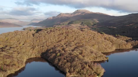 Tomas-De-Drones-Ascendentes-Y-Panorámicos-De-Un-Amanecer-De-Otoño-Sobre-Loch-Lomond-Y-La-Montaña-Ben-Lomond-En-El-Parque-Nacional-Loch-Lomond-Y-Los-Trossachs,-Escocia-Con-Bosques-Nativos-De-Hoja-Ancha-En-La-Orilla