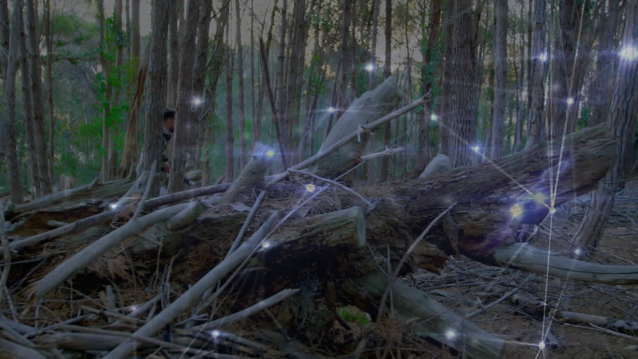 Man crouching behind logs in pine forest, showing rifle and digital sparkles for defense simulation