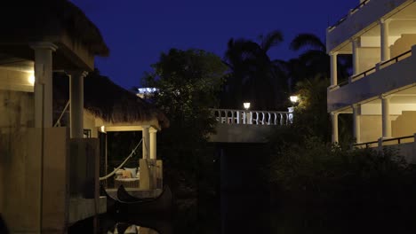 tranquil night time scene of small tropical bungalow's along a small river with a car driving by on the illuminated bridge above in a vacation resort in riviera maya, mexico near cancun and tulum