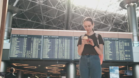 young beautiful woman traveler standing at the airport with a backpack and looking at the smartphone screen