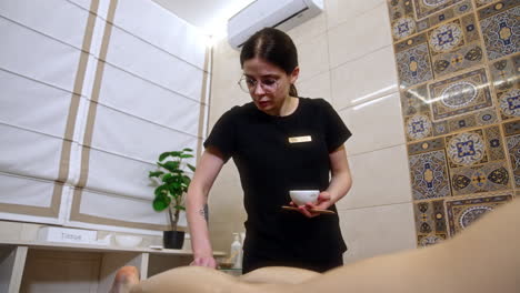 thin dark-haired woman covers the client's legs with cream. cosmetologist holding a cup with cream in her hand.