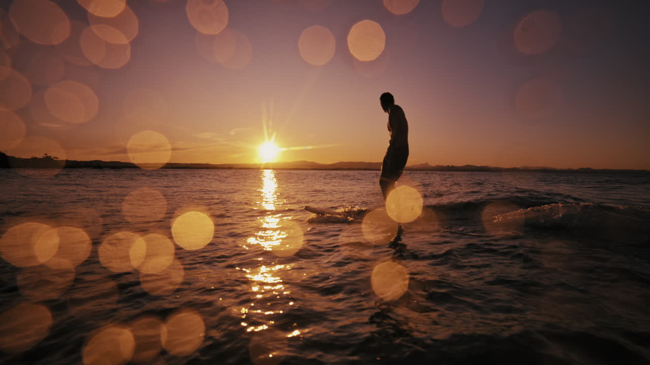 Surfer walks by glowing water at sunset with soft light circles in foreground, longboard cruising, slow motion silhouette