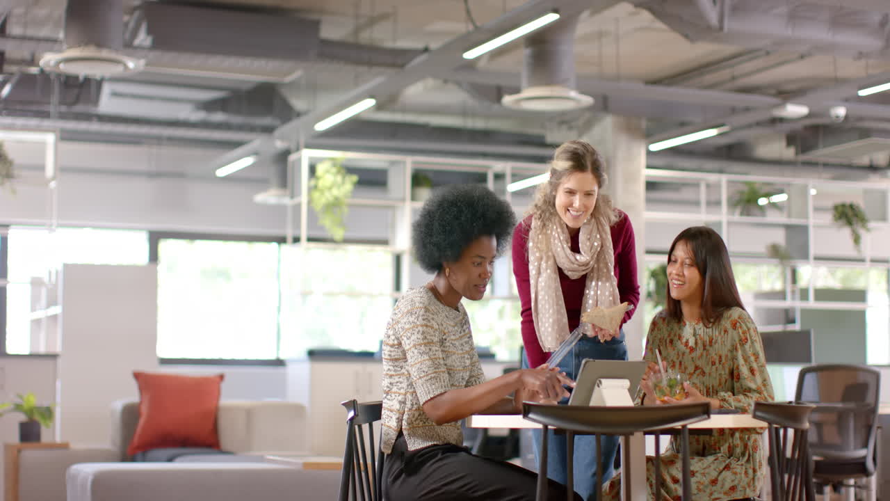Diverse female creative colleagues discussing using tablet and eating lunch in office, slow motion