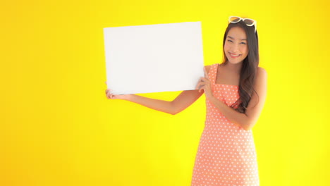 young asian woman in summer dress with sunglasses on head holds up plain white sign board and smiles