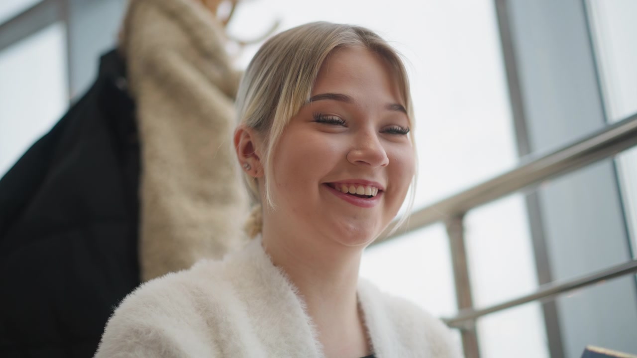 Young blonde woman seated by large bright window smiles with lifted cheeks and subtle head tilt, suggesting relaxed attentive conversation, quiet amusement, and calm friendly indoor atmosphere