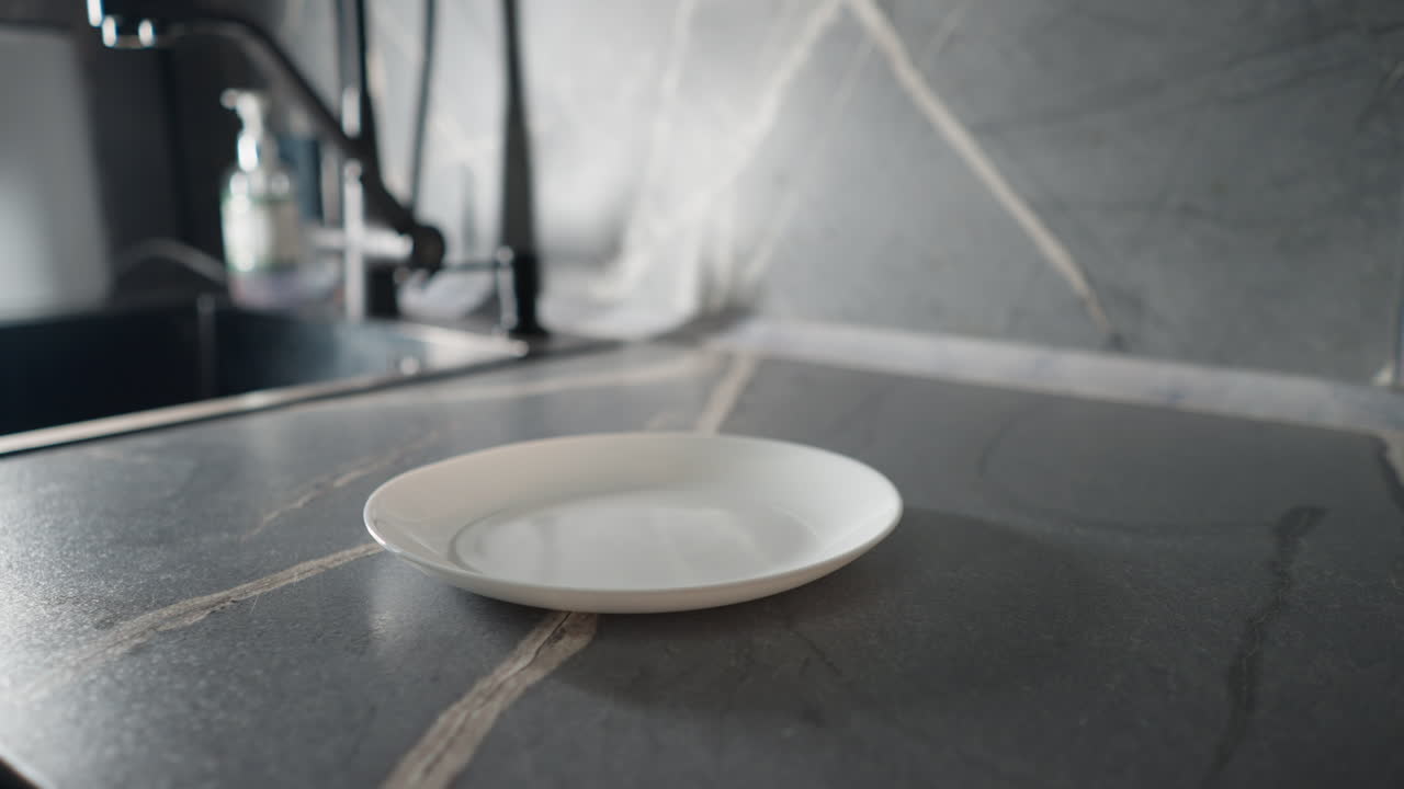 Elderly woman in green sweatshirt placing food on empty white plate in modern kitchen. Focus on hands and plate as part of her daily cooking and meal preparation routine