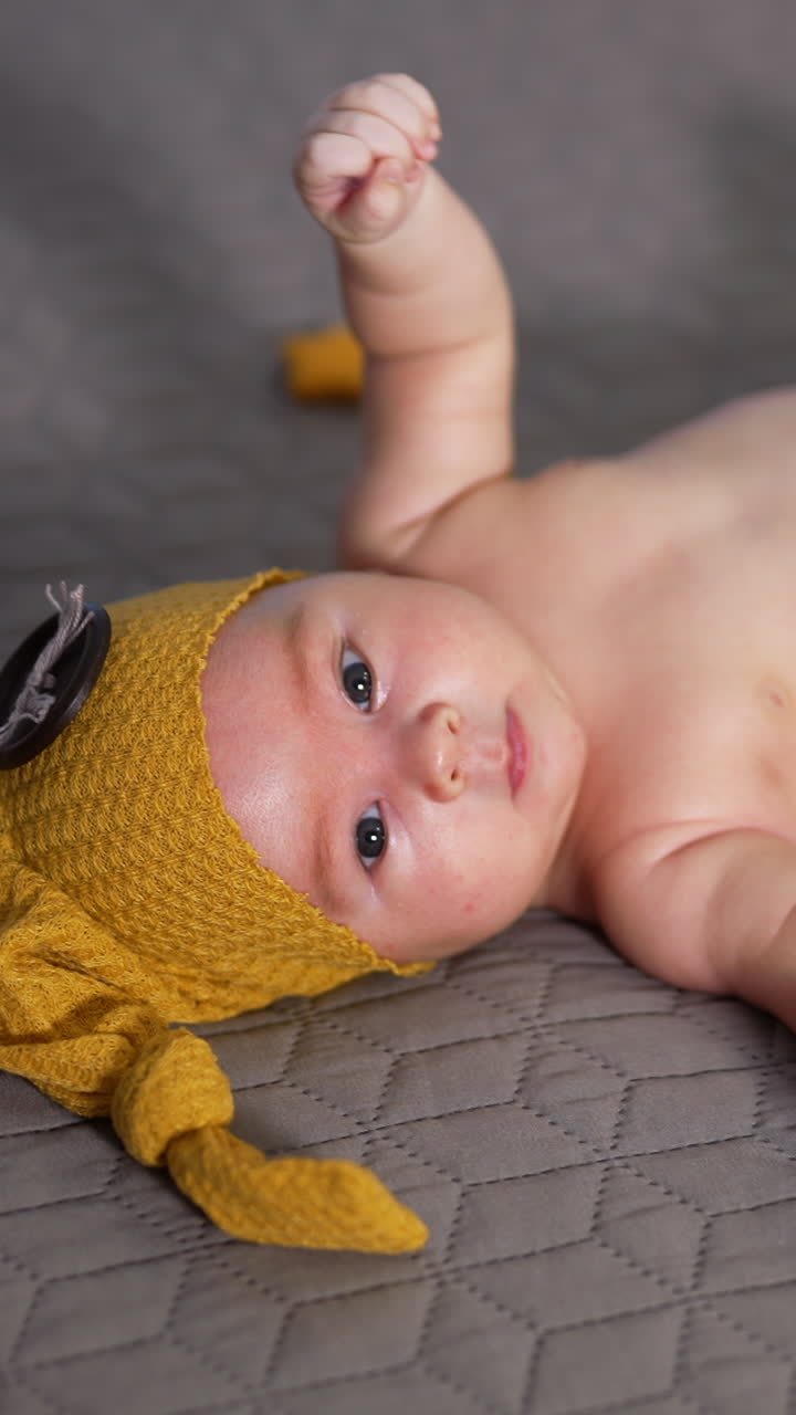 Small boy waving his arms and feet while lying on the bed. Boy in yellow costume with bare belly. Kid in a funny hat with big button. Vertical video