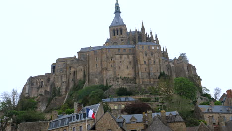 Tiro-Inclinado-Hacia-La-Catedral-De-Le-Mont-Saint-Michel-Y-Ondeando-Banderas-Francesas-Contra-El-Cielo-Nublado