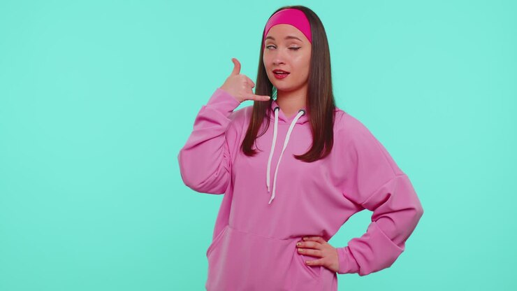 Ragazza adolescente con felpa rosa con cappuccio che guarda in camera e fa il gesto del telefono, come a dire "ehi, richiamami".