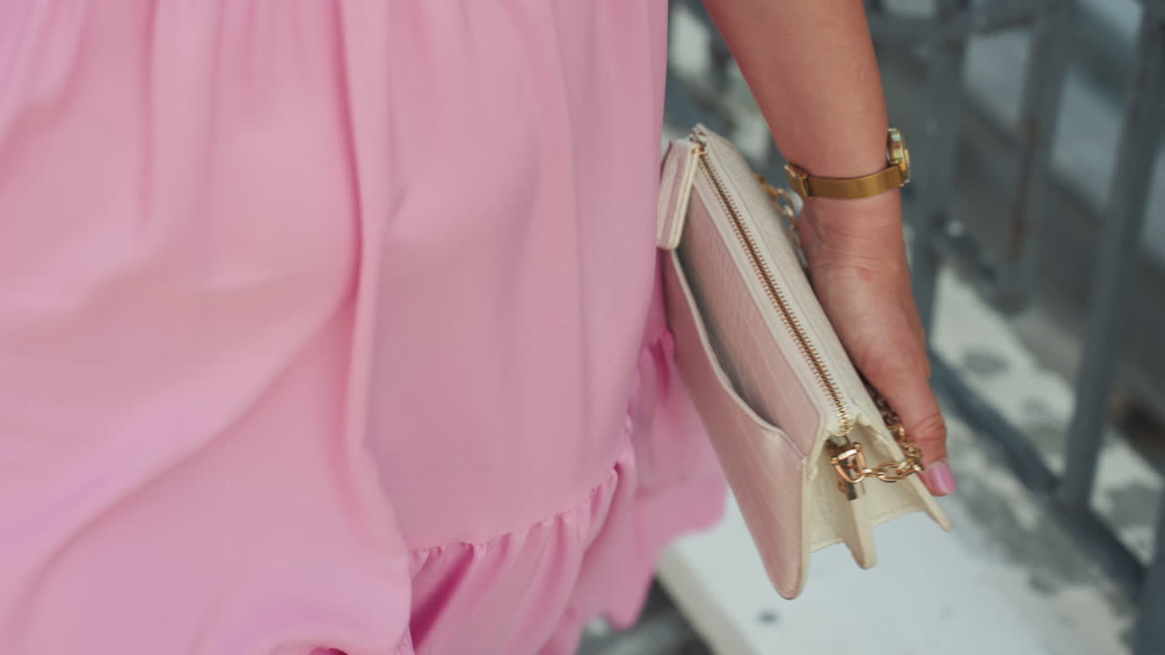 Donna bianca con abito rosa che tiene in mano una pochette, in piedi accanto a una ringhiera in ferro, pochette color oro rosa e braccialetto dorato, orlo a volantFluidi, luce diurna soffusa, texture del tessuto tattile, atmosfera editoriale romantica, femminile