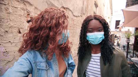 caucasian and african american women wearing masks and waving at camera while smiling in the street