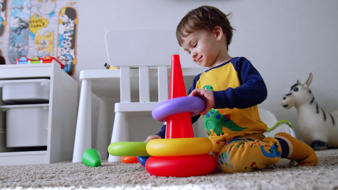 Cute baby boy sits down on the floor near the toy pyramid. Kid assembles the toy cheerfully. Low angle view.