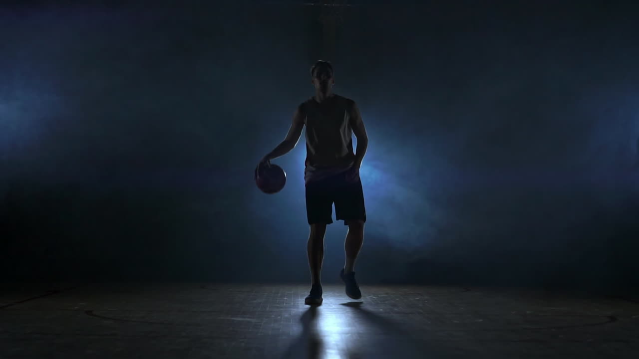 el jugador de baloncesto va directamente a la cámara en una habitación oscura con una luz de fondo en el humo mirando a la cámara en cámara lenta. steadicam