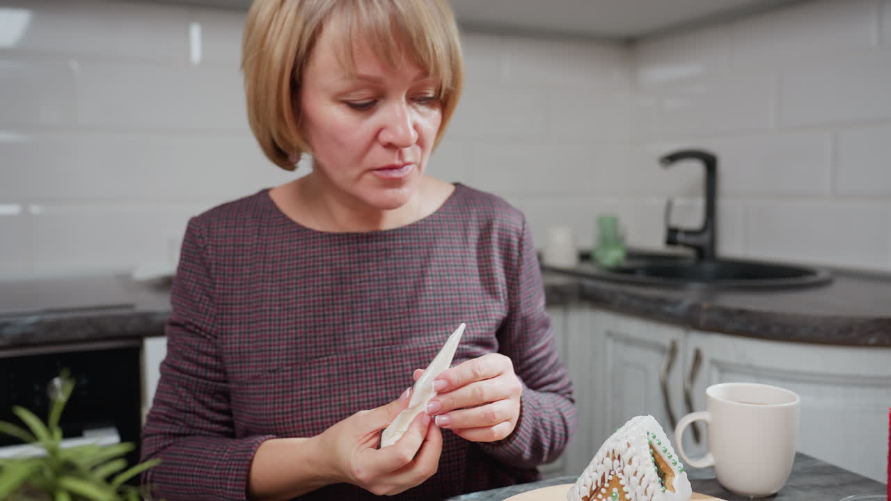 mujer de mediana edad sosteniendo una bolsa de helado, decorando cuidadosamente la casa de pan de jengibre en una cocina cálida, vela, taza, galletas y planta en olla en la mesa
