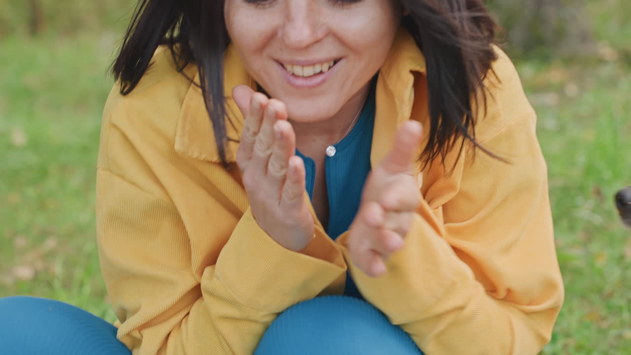 Donna che si scalda le mani al campeggio, seduta sull'erba, con una giacca gialla e occhiali da sole sulla testa. Un sorriso delicato, espirando fiato caldo, in un momento di campeggio intimo e accogliente in un bosco di betulle, un primo piano spontaneo.