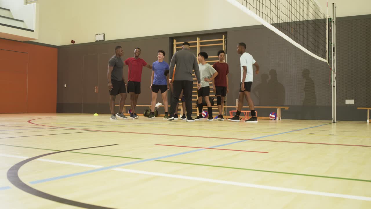 Mangfoldigt mandligt volleyballhold i spilledragter, der lytter til træneren, der klapper, mens de samler volleyballbolde ved nettet i gymnastiksalen.