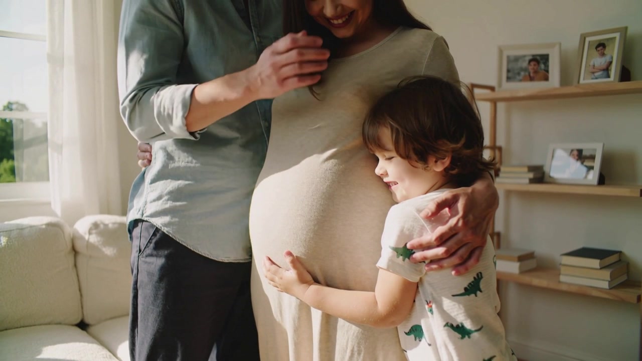 Bambino che tocca il pancione della mamma in abito beige, mentre il padre la abbraccia, creando un legame affettuoso vicino alla finestra.
