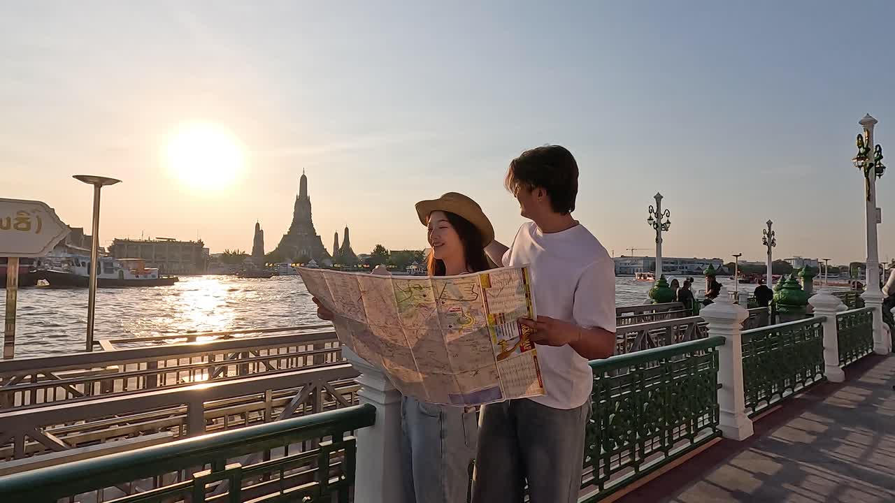 Coppia di turisti romantica studia una mappa cartacea su una passeggiata lungofiume all'ora d'oro, con un tempio sullo sfondo.