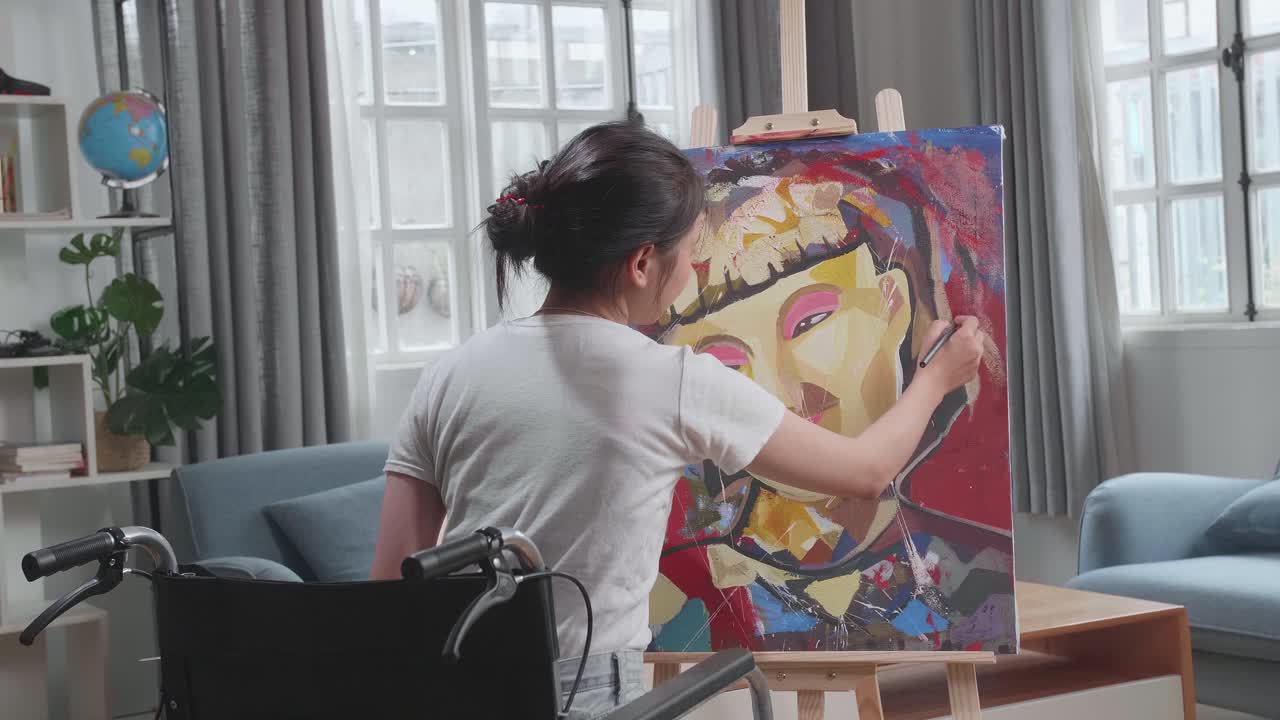 Back View Of An Asian Artist Girl In Wheelchair Holding Paintbrush Mixed Colour And Painting A Girl On The Canvas