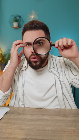 Freelancer-man-holds-magnifying-glass-near-face-looking-at-camera-with-big-zoomed-eye,-analyzing