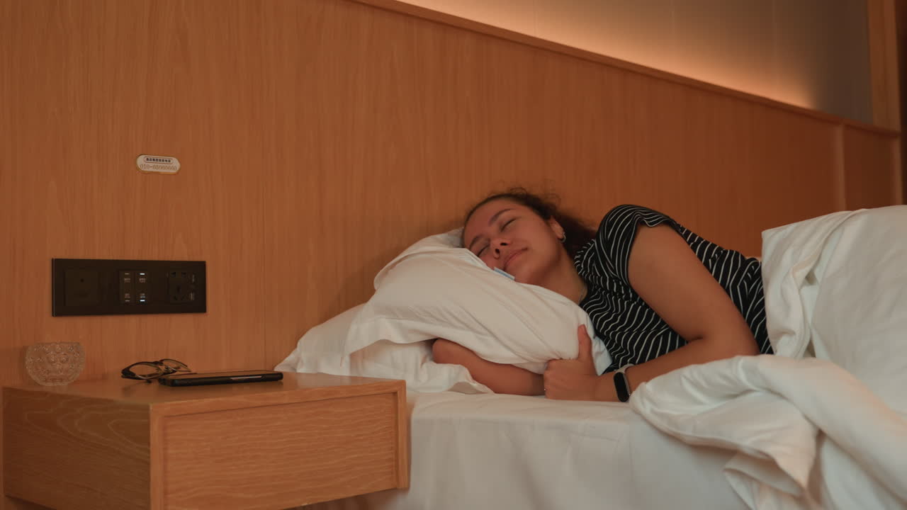 Solsmukt hotelværelse, fredfyldt kvinde hviler på puden, med et mildt smil og afslappet kropsholdning, blødt hvidt sengetøj, varm træsengegavl, rolig morgenstemning med gæstfrihedsdetaljer, en følelse af lettelse efter rejsen.