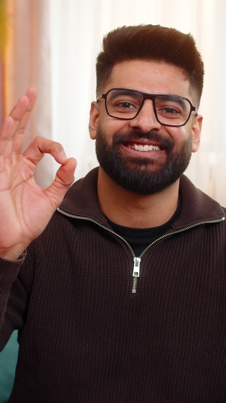 Indian man on sofa smiles and shows ok gesture to camera at home with confident positive mood