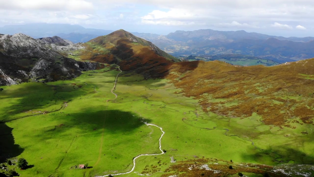 Scena dall'alto su una vasta valle montana con pascoli di un verde vivido, un sentiero pallido e tortuoso, un ruscello stretto, colline marrone ruggine, ombre soffici delle nuvole, lievi cambiamenti di luce, un'ambientazione di viaggio serena.