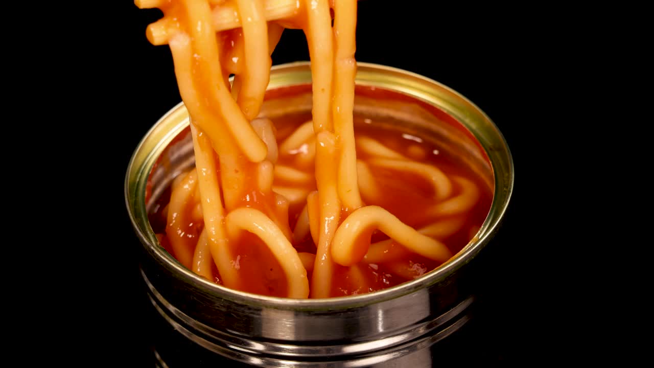 Chopsticks grasp and lift spaghetti noodles from an open metal can filled with tomato sauce, shot against a black background with even studio lighting