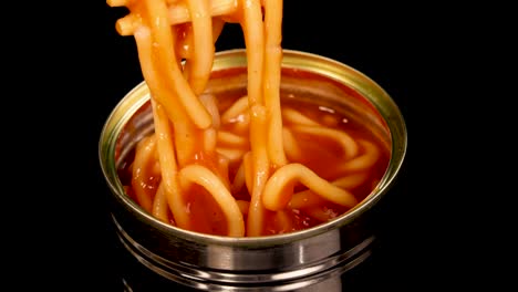 chopsticks grasp and lift spaghetti noodles from an open metal can filled with tomato sauce, shot against a black background with even studio lighting