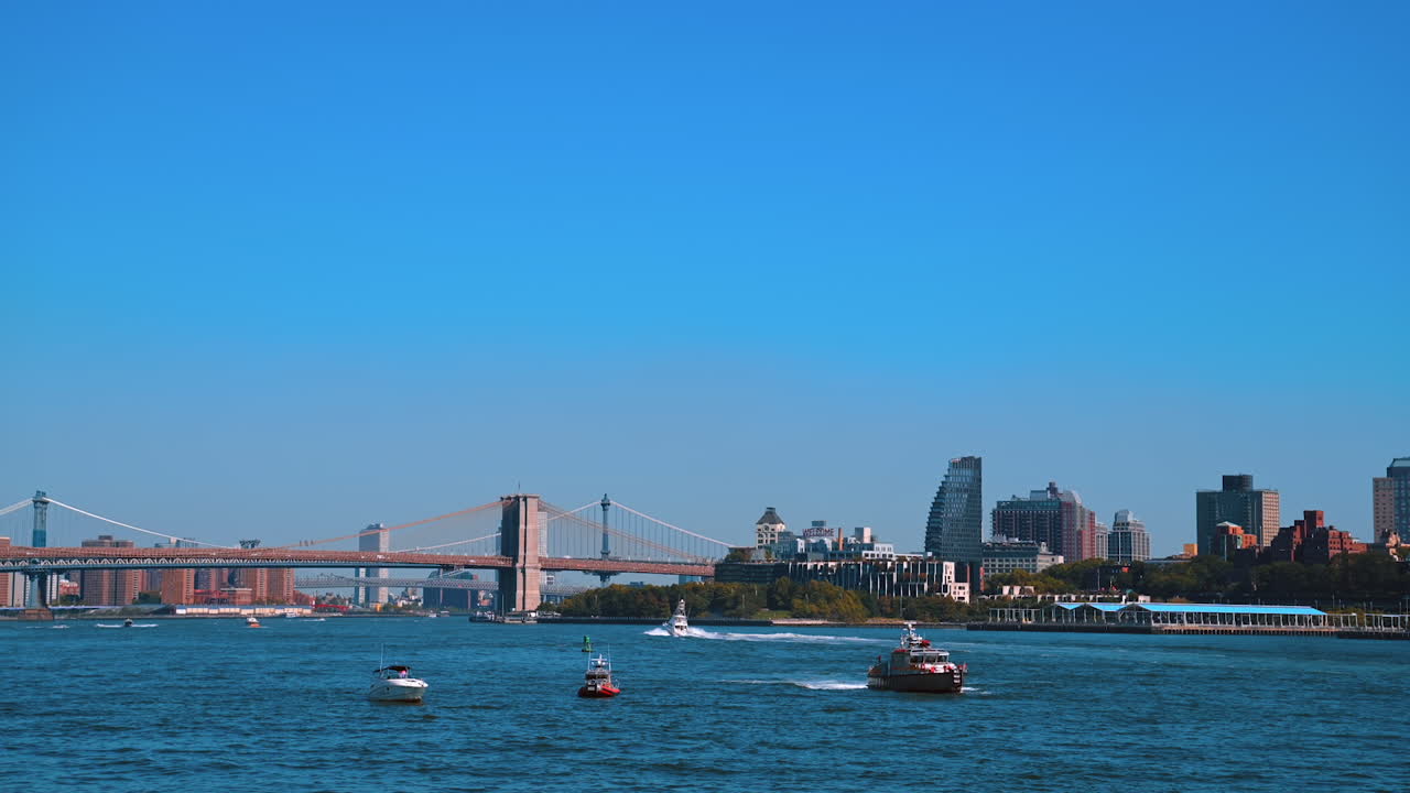 Many different boats travel by the east River on sunny day. View from the riverscape. The Brooklyn Bridge and Manhattan Bridge and city skyline at backdrop