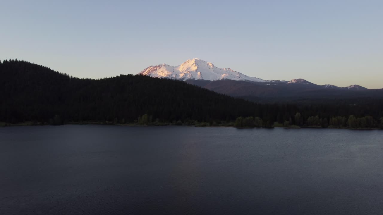 Sunset counter-clockwise reveal drone shot of Mount Shasta volcano at sunset. The colors of the sunset reflect on the water of Lake Siskiyou as the glacier-covered mountain peaks from behind the hill.