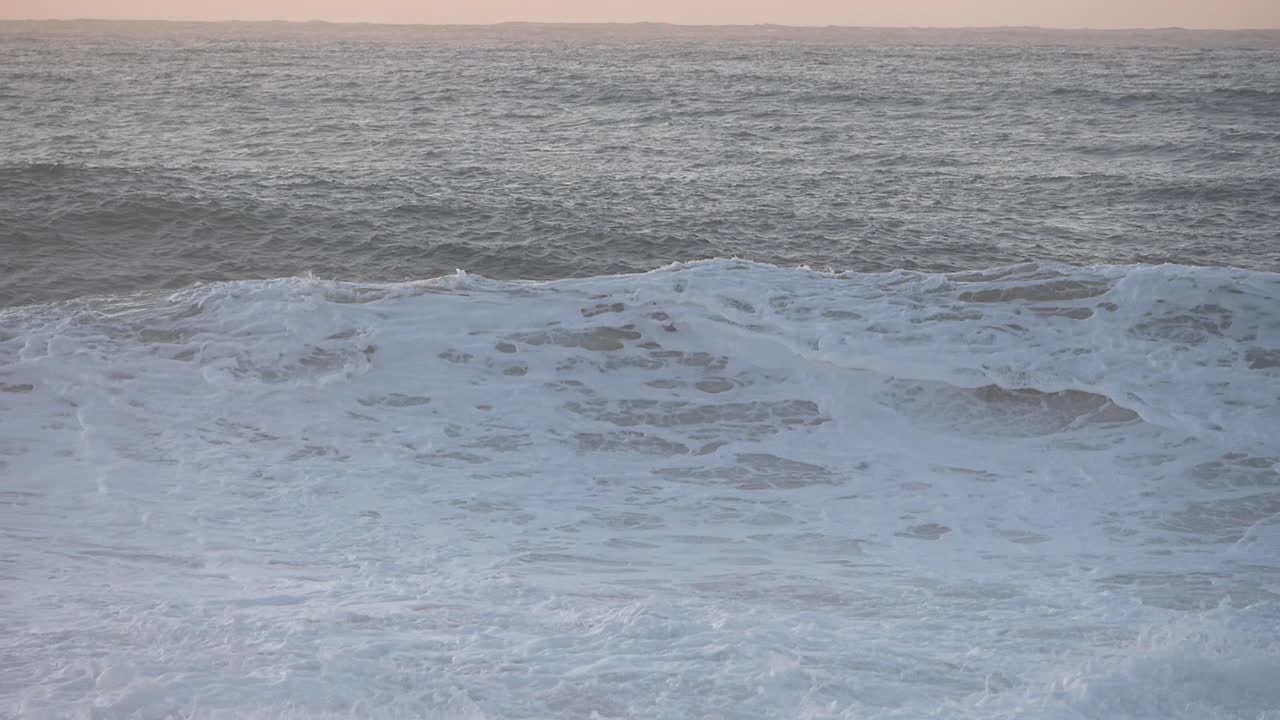 Close-up of big waves at beachfront crashing during sunset. Golden light on horizon. Tall waves curl and break, big splashes. Wind blows water  particles from wave crest.
