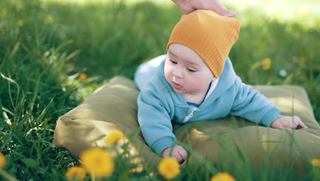 little child in blue overall and orange cap looks around in the nature. mother's hand carefully pulls son's hat.
