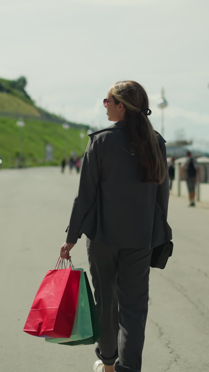 회색 옷을 입은 여인이 빨간색, 민트색, 녹색 쇼핑 가방을 들고 다리 위를 고 있는 모습.