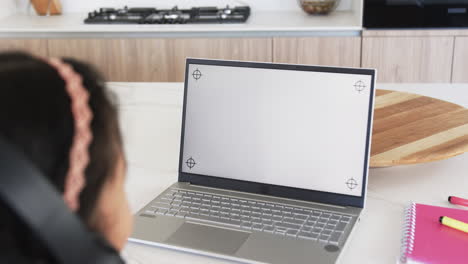 Biracial-girl-with-braided-hair-is-using-a-laptop-in-a-modern-kitchen,-with-copy-space