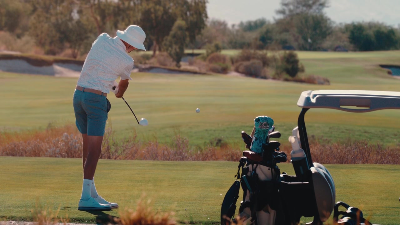 Golfer hits 3 wood off the tee, sending the ball toward distant desert mountains in warm golden light, captured in slow motion