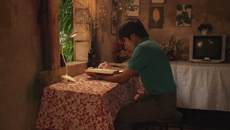 young asian male reads a book at a desk in a rustic room