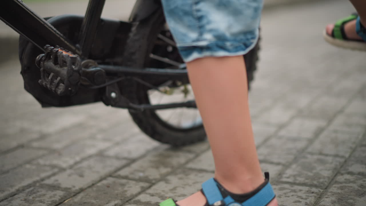 그는 자전거 자전거를 움직이고 타고 다니면서 다채로운 샌달과 파란색 청바지 쇼트를 입은 아이의 다리의 클로즈업