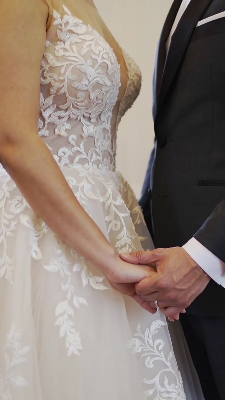 Vertical video: Holding bride in lace dress, groom in suit holding hands during vows, showing bands