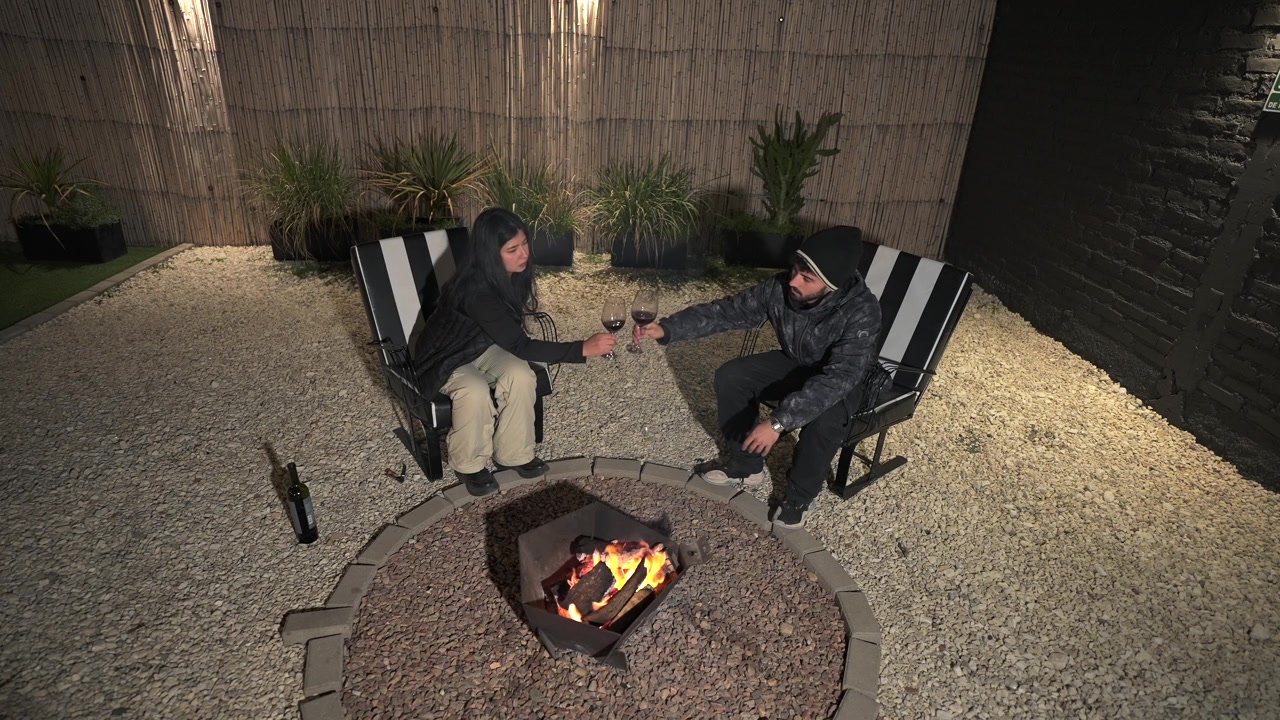 Coppia che brinda con bicchieri di vino rosso accanto a un braciere da esterno su un patio privato nel cortile, godendosi la conversazione e il relax in un'atmosfera serale accogliente, vista statica dall'alto