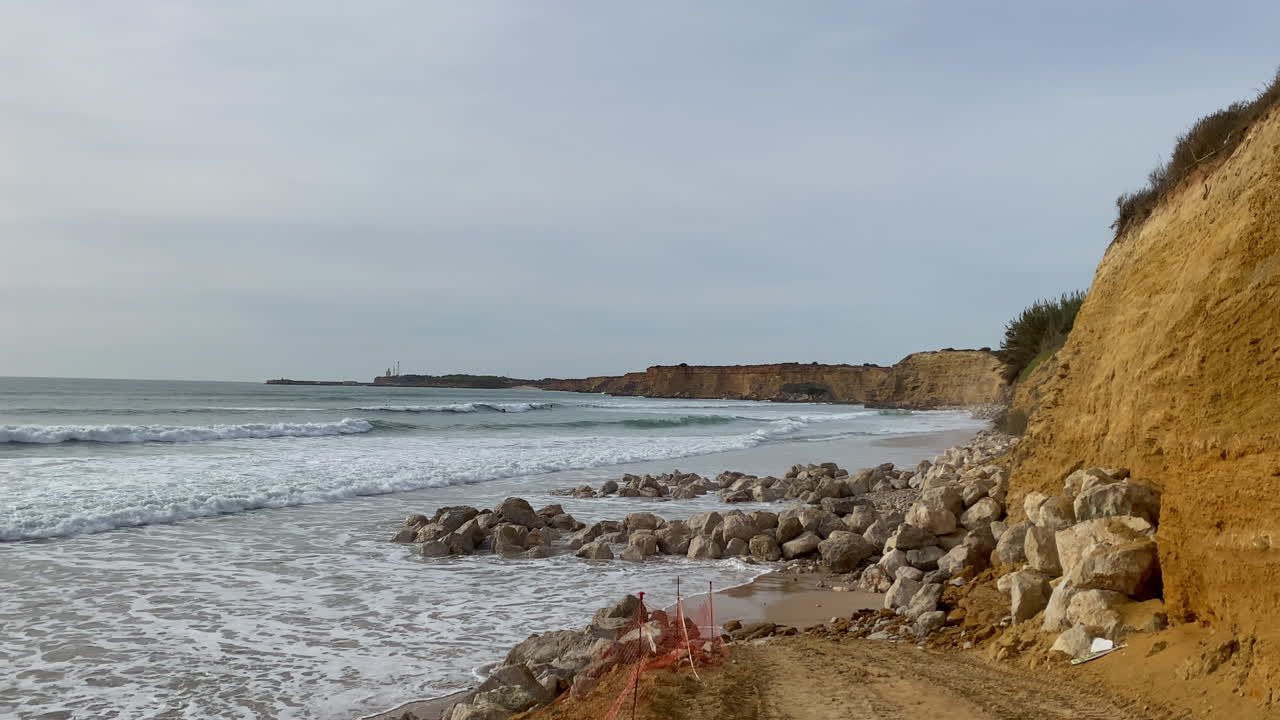 Sea defences of piled rocks protect the eroding cliffs while waves push onto the narrow winter beach at Fuente del Gallo